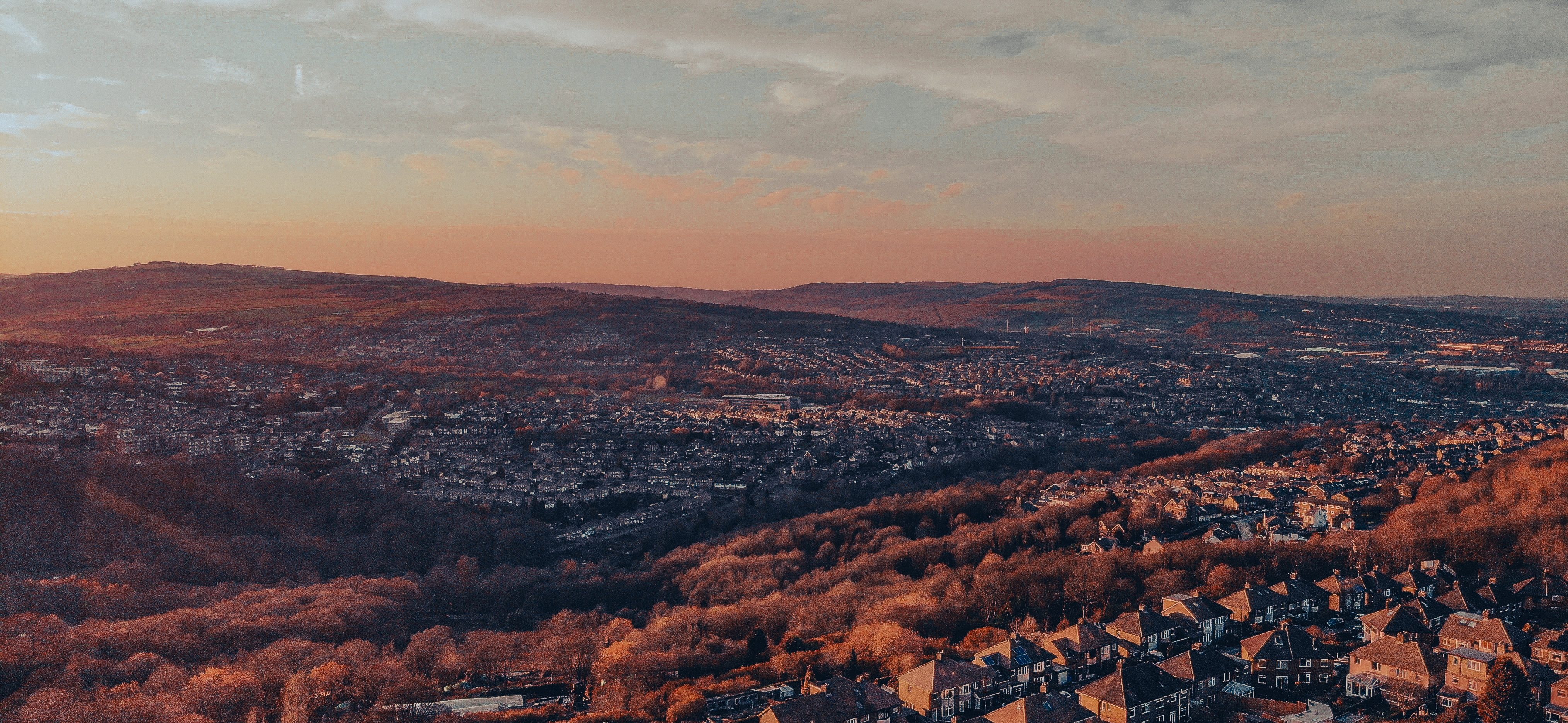 Aerial view of a scenic landscape at sunset in Sheffield, featuring a town surrounded by rolling hills and dense forests. The sky is painted with warm hues of orange and pink, casting a soft glow over the rooftops; an inviting scene perfect for students seeking inspiration for things to do outdoors.