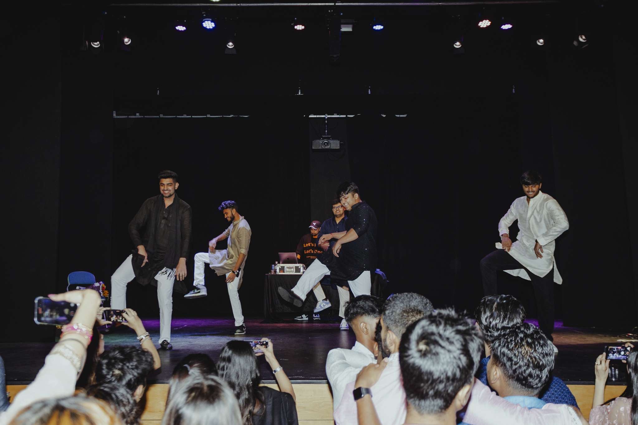 A group of men performs a traditional dance on stage, wearing vibrant attire. The university audience watches and records with phones. In the background, a DJ stands at a table with equipment under colorful stage lights, guiding the rhythm and energy of the evening.