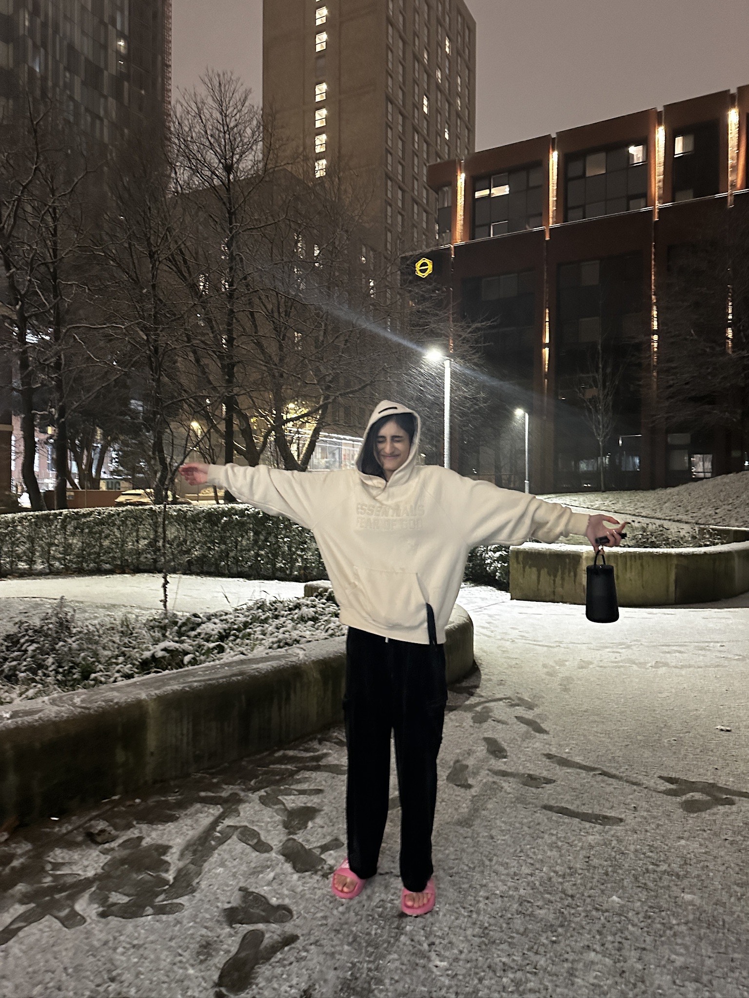 A university student stands outside at night in a snowy urban area, wearing a hoodie and sandals while clutching a water bottle. Arms outstretched, they're surrounded by tall buildings and bare trees—perhaps contemplating the best student advice as they walk along the snow-dusted pathway.