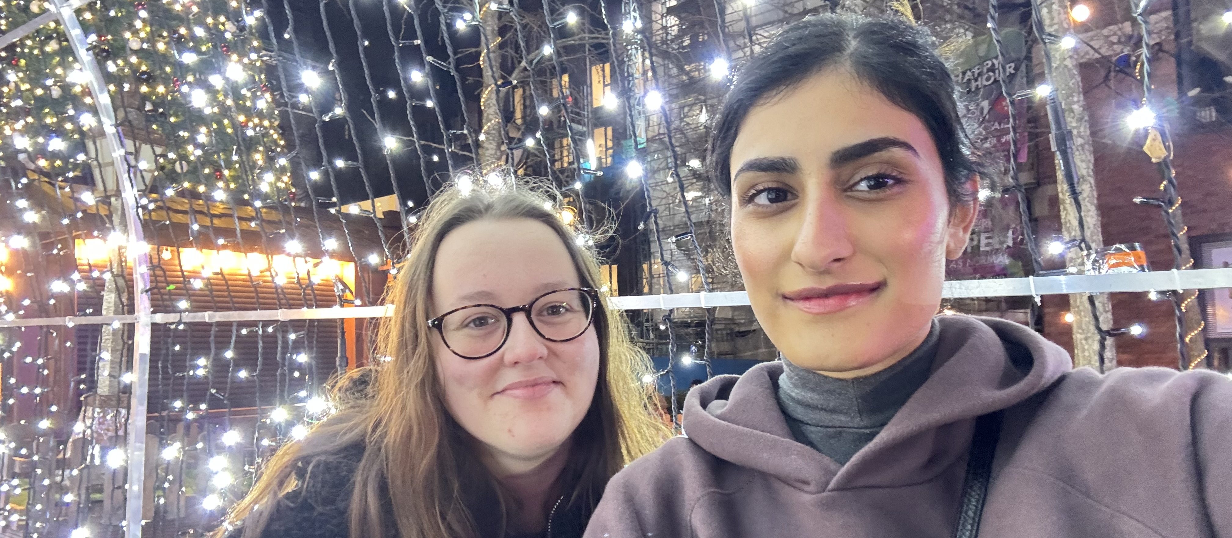 Two people are smiling and taking a selfie in front of a festive backdrop adorned with bright white lights and illuminated trees. The area is dimly lit, enhancing the joyful atmosphere—a perfect snapshot of the vibrant resident experience at Vita Student, where making friends comes naturally.