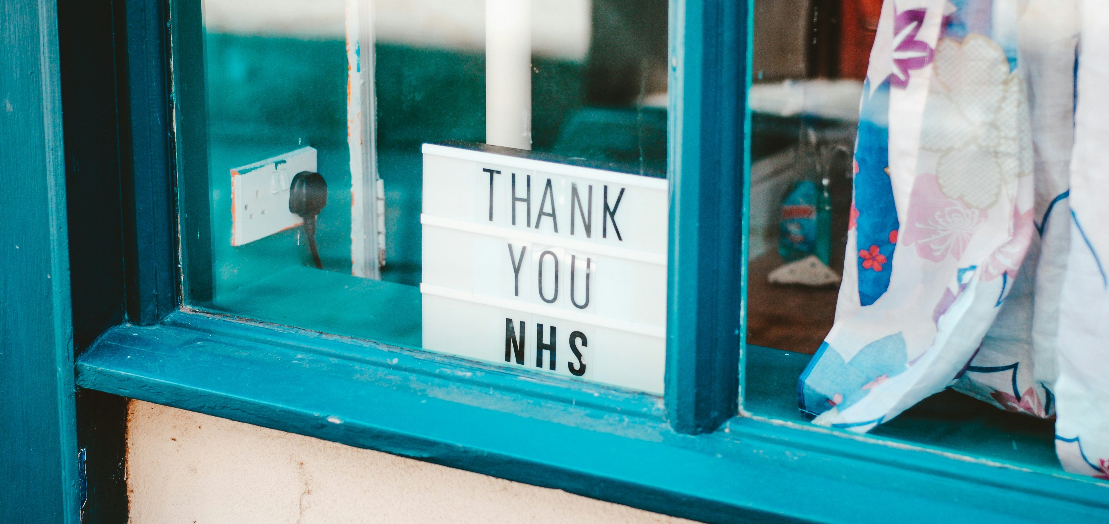 A lightbox in a window displays the message "Thank You NHS," celebrating the vital role of healthcare for international students in the UK. The window frame is painted blue, adorned with colorful floral fabric nearby, highlighting the community's gratitude.
