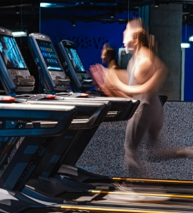 A person in motion is running on a treadmill at a gym, their speed captured with a blurred effect. Nearby, friends cheer them on, reflected subtly in the mirror. Several treadmills are lined up in the modern, dark interior, making it an ideal spot to refer others for an energizing workout.