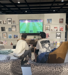 Two friends sit on a cozy living room couch, engrossed in a soccer video game on a large wall-mounted TV. The wall behind them showcases artworks and photographs. As they play, they talk about the exciting Friend Rewards they'll get when they refer your friends to their favorite gaming platform.