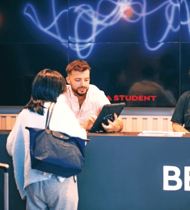 A man is seated behind a counter, engaging with a woman holding a tablet. The woman, wearing a backpack, stands facing the counter. The background features a large screen with an abstract design and text that includes "STUDENT." It seems they refer to friends about the academic event advertised.