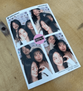 A sheet of photobooth images featuring three friends posing together with playful expressions. The background has a repeating pattern and text saying "CHOW DOWN" in the center, a nod to their shared adventures. The sheet rests on a wooden surface.