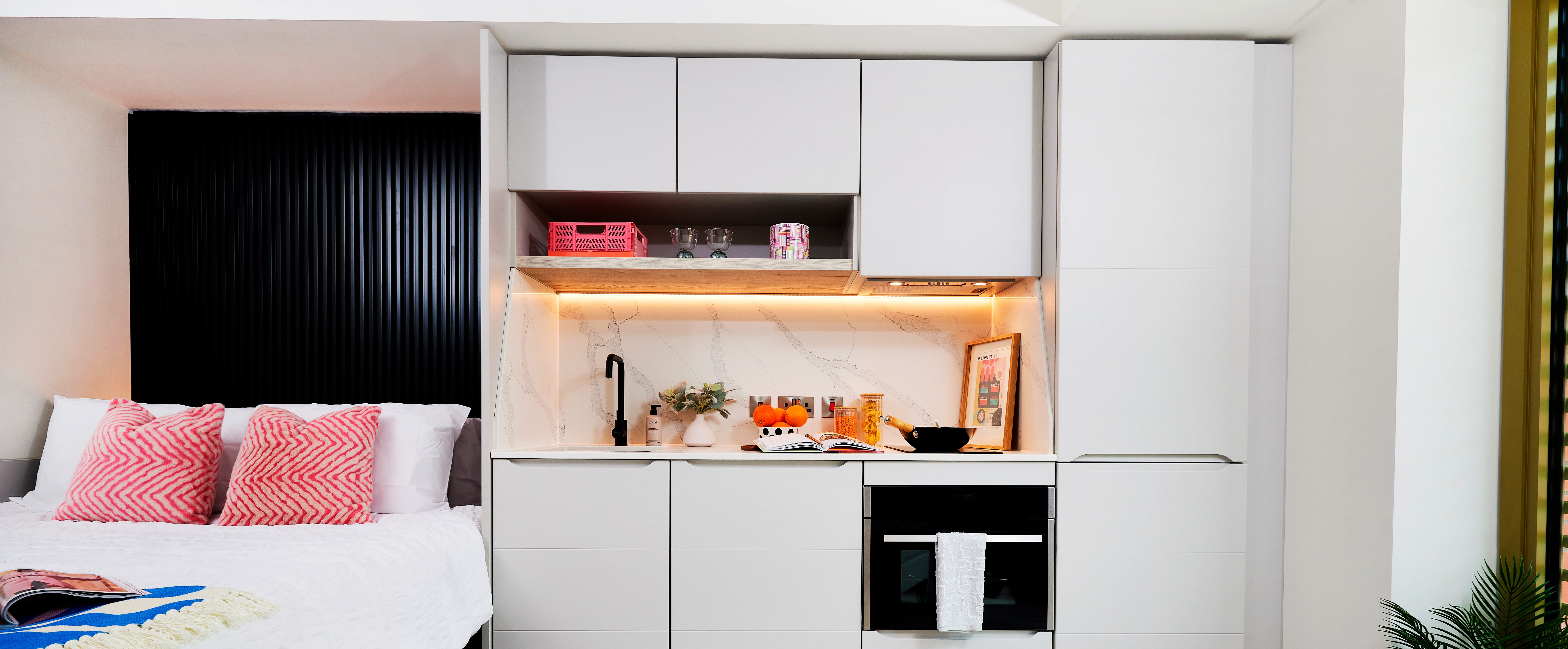 This compact modern studio apartment is ideal for student accommodation. It features a small kitchenette with white cabinets, a black faucet, and an oven. The countertop has decor items, while soft pink lighting accents the area. A bed with pink pillows is adjacent to the kitchen.