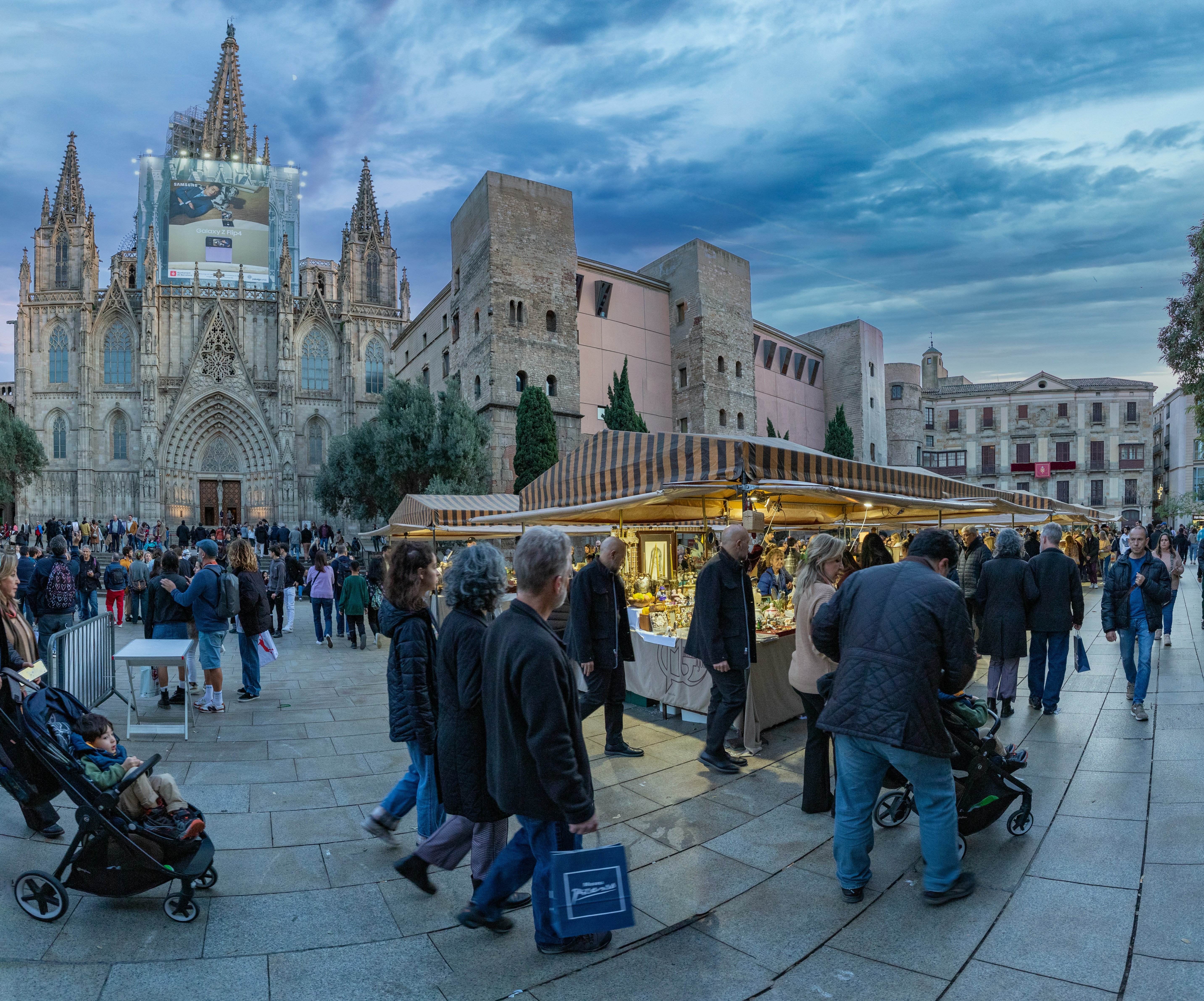 People stroll through a bustling outdoor market in front of a large Gothic cathedral with a circular window. The lively scene, set under a cloudy invierno sky, is filled with vendor stalls and clusters of estudiantes exploring the vibrant atmosphere in the heart of Barcelona.