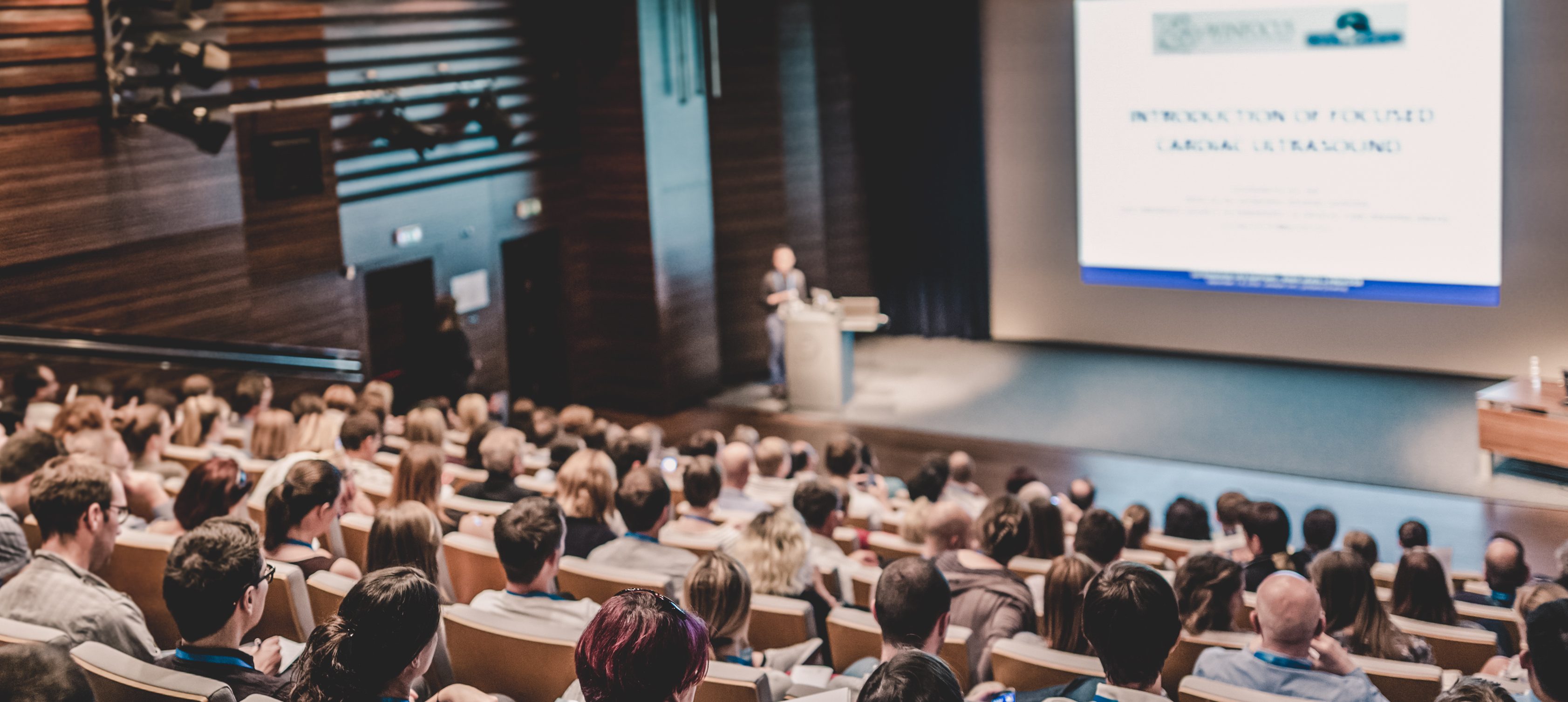 A large audience is seated in a lecture hall, including many eager second-year students, listening to a presentation. A speaker stands at a podium in front of a screen with a slide titled "INTRODUCTION OF FOCUSED (VIRTUAL TRACKING)," offering valuable tips. The room features wooden walls and a modern design.