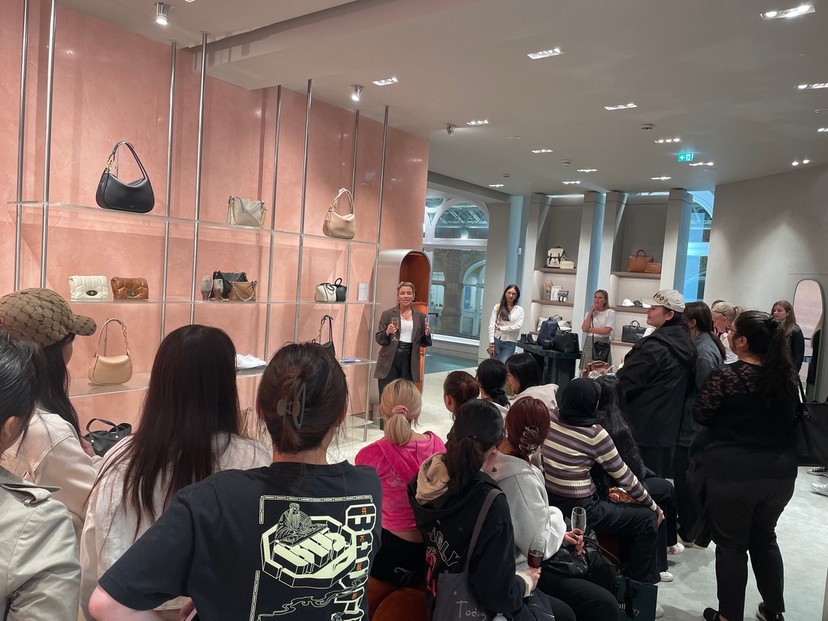 A group of people, eager to explore the latest Vita Student trends, gather in a store with handbags displayed on the shelves. A brand partner stands in front, speaking to the attentive audience. The room is well-lit, and some attendees are seated while others stand.