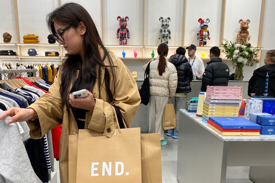 A woman with long hair and glasses browses clothing in a store. Wearing a tan coat, she holds a "Vita Student" shopping bag. Art toys and decorative items are displayed on shelves in the background, where several people are at the checkout counter.