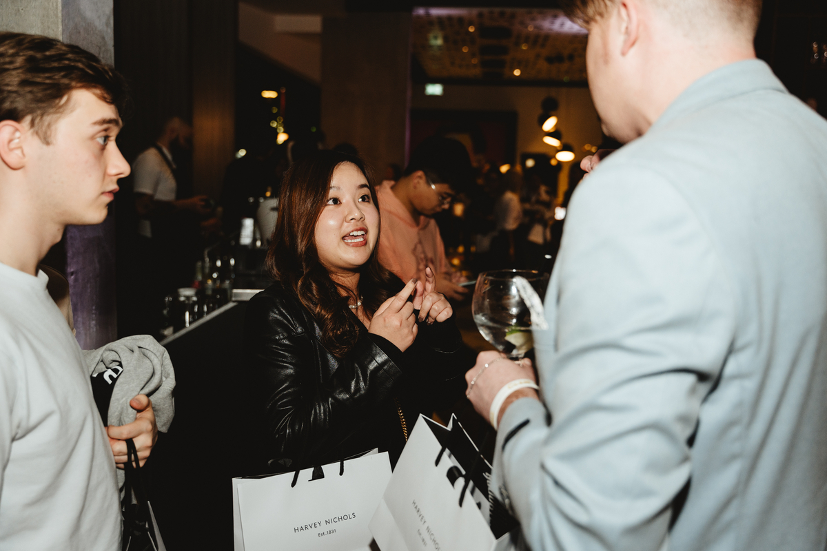 A woman in a black jacket, perhaps a Vita Student Brand Partner, gestures animatedly while chatting with a man in a light blue jacket. Both clutch Harvey Nichols shopping bags. Nearby, another man listens attentively amidst the dim lighting and mingling crowd.