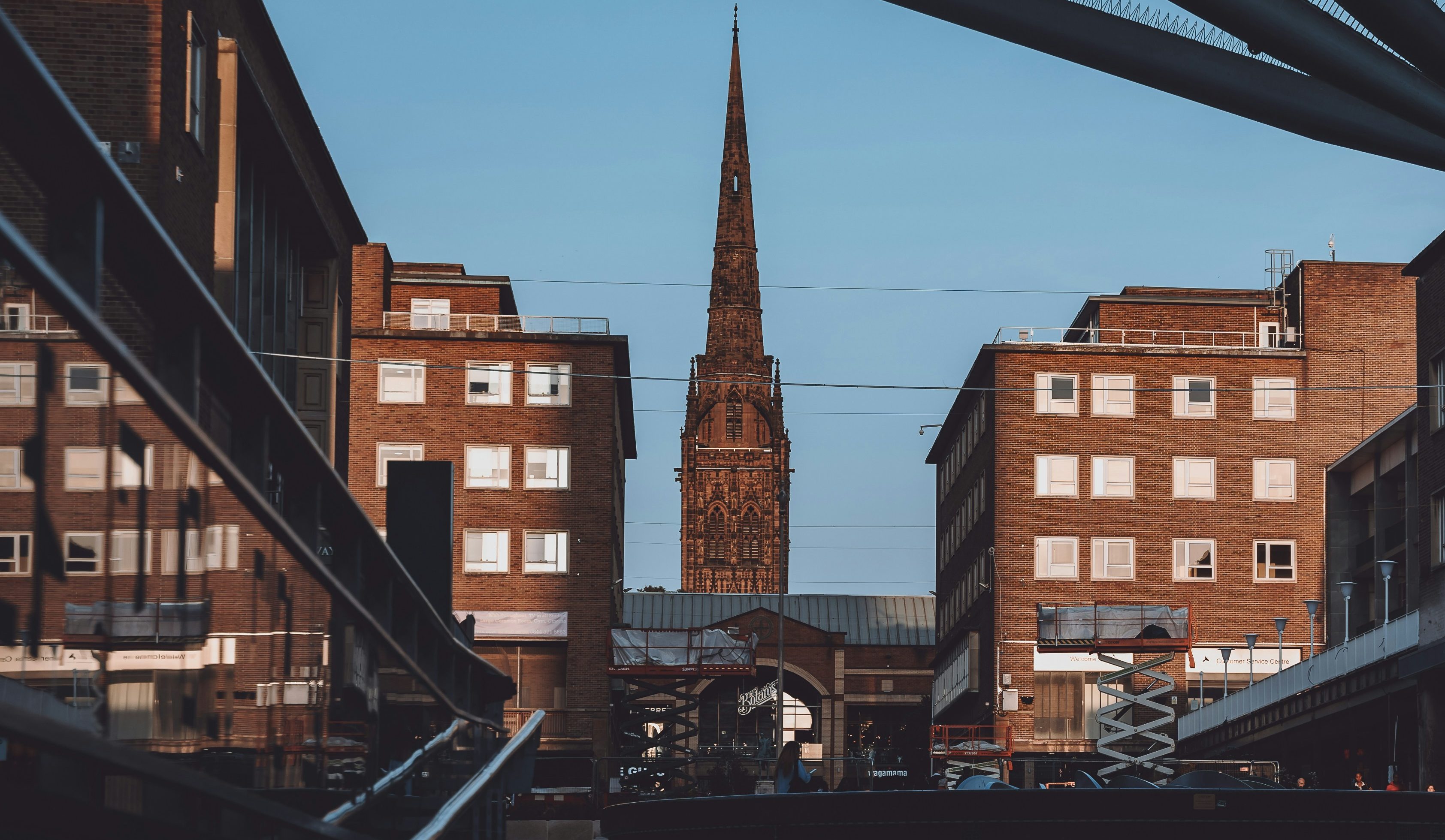 A tall, pointed church spire is centered between brown brick buildings under a clear blue sky, reminiscent of heritage sights for anyone relocating to Coventry. The scene includes reflections on nearby glass surfaces, with construction equipment visible in the lower foreground.