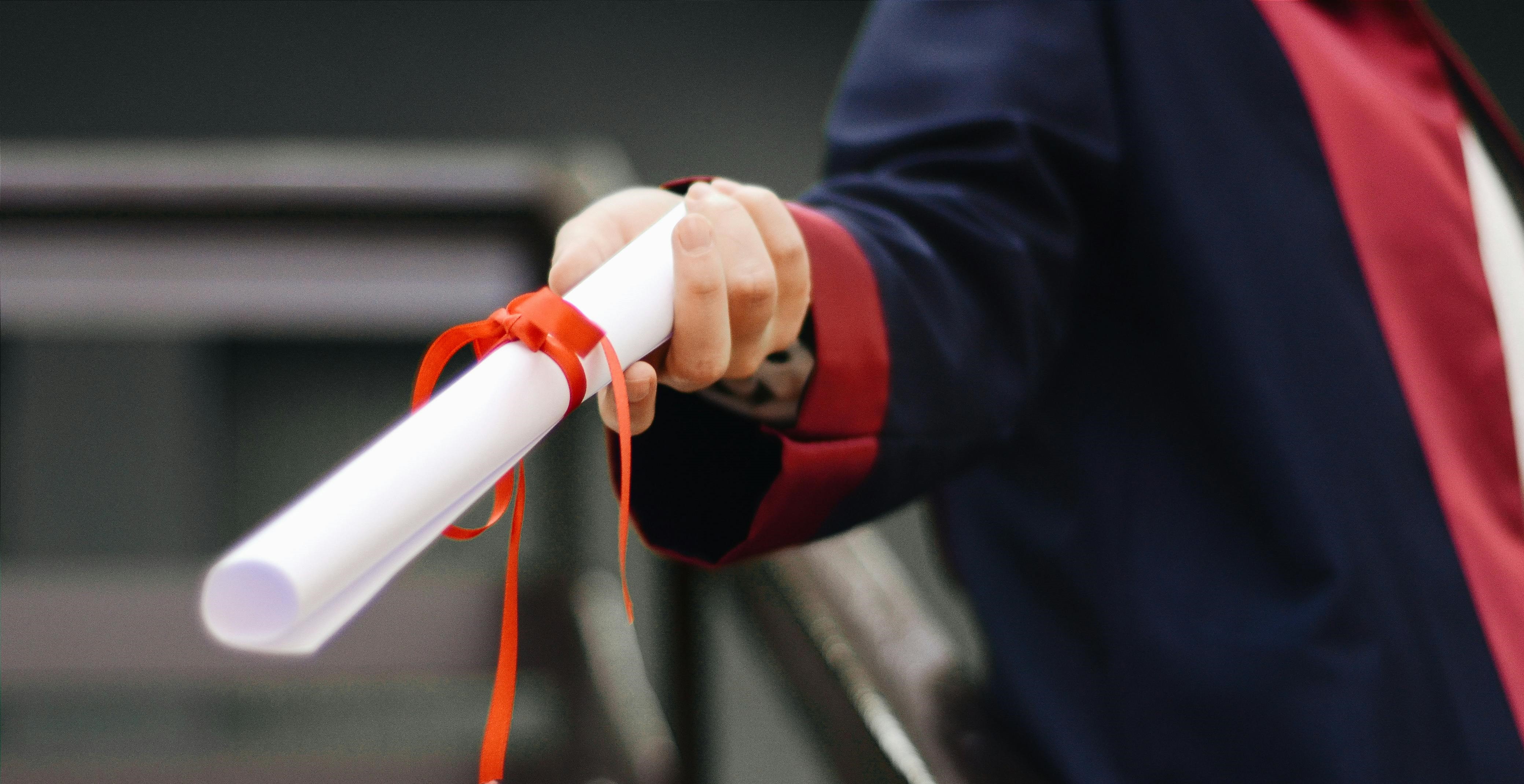 A person wearing a dark graduation gown and red sash proudly holds out a rolled diploma tied with a red ribbon. The focus is on the hand and diploma, symbolizing the journey to homologar their título extranjero in España. The background remains elegantly blurred.