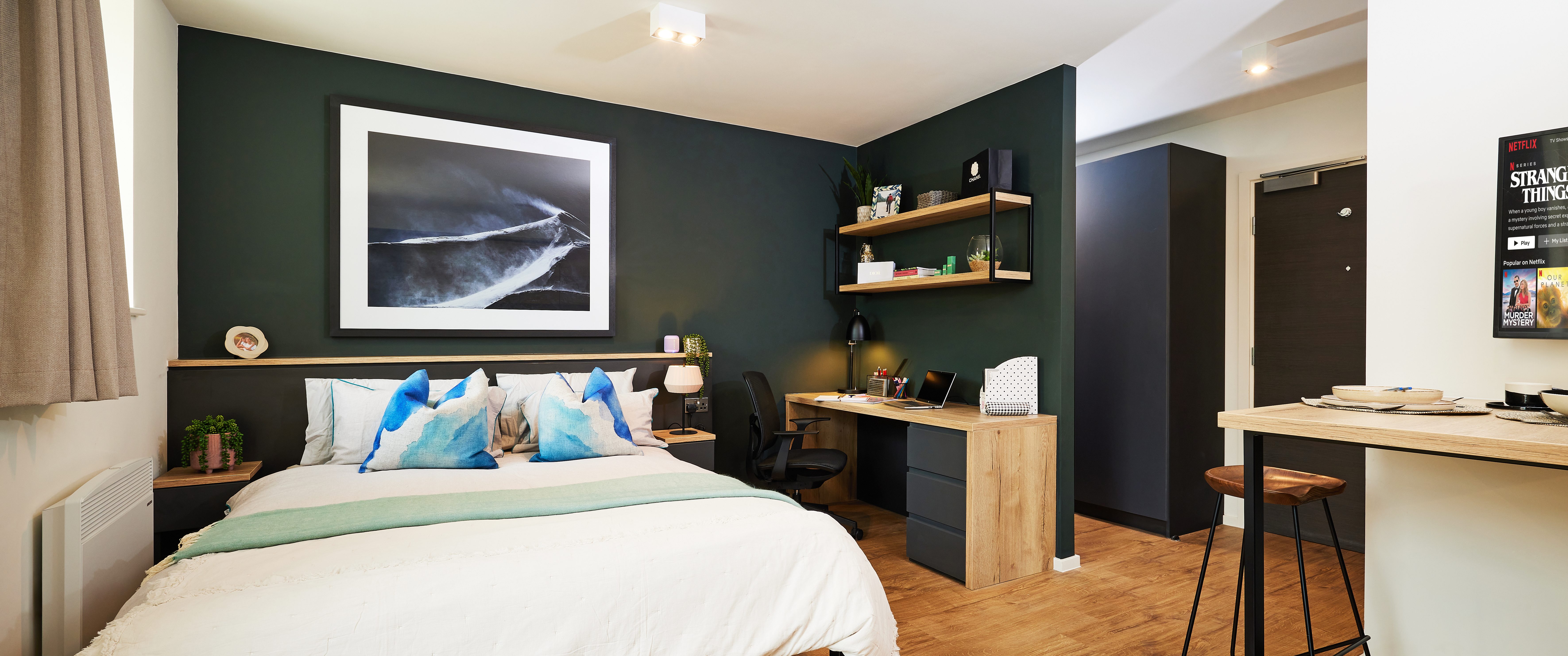 This modern student accommodation features a spacious bedroom with a large bed, white and teal pillows, and a soft green blanket. A dark accent wall showcases a framed abstract photo. Wooden shelves above the desk hold plants and books, while light wood floors and wall art create a cozy atmosphere.
