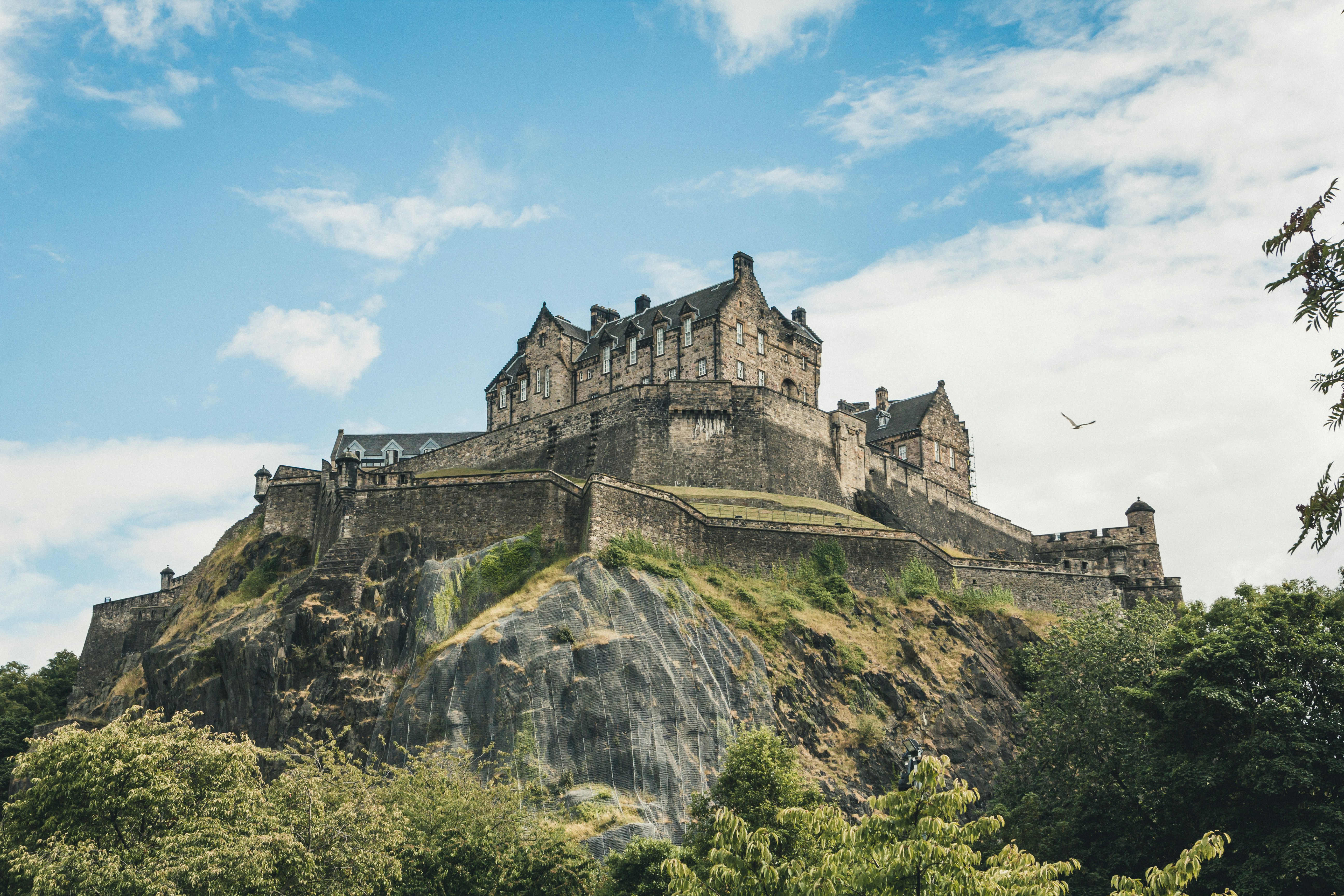 Stone castle perched on a rocky hill, surrounded by green trees under a partly cloudy sky. A single bird flies nearby, enhancing the picturesque and historic atmosphere of Explora Edimburgo.