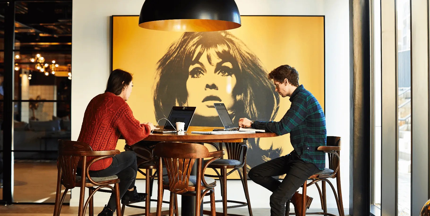 Two people work on laptops at a wooden table in a modern cafe. They sit beneath a large black lamp, with a bold, black-and-yellow artwork of a woman's face in the background. The space has a warm light and contemporary design.