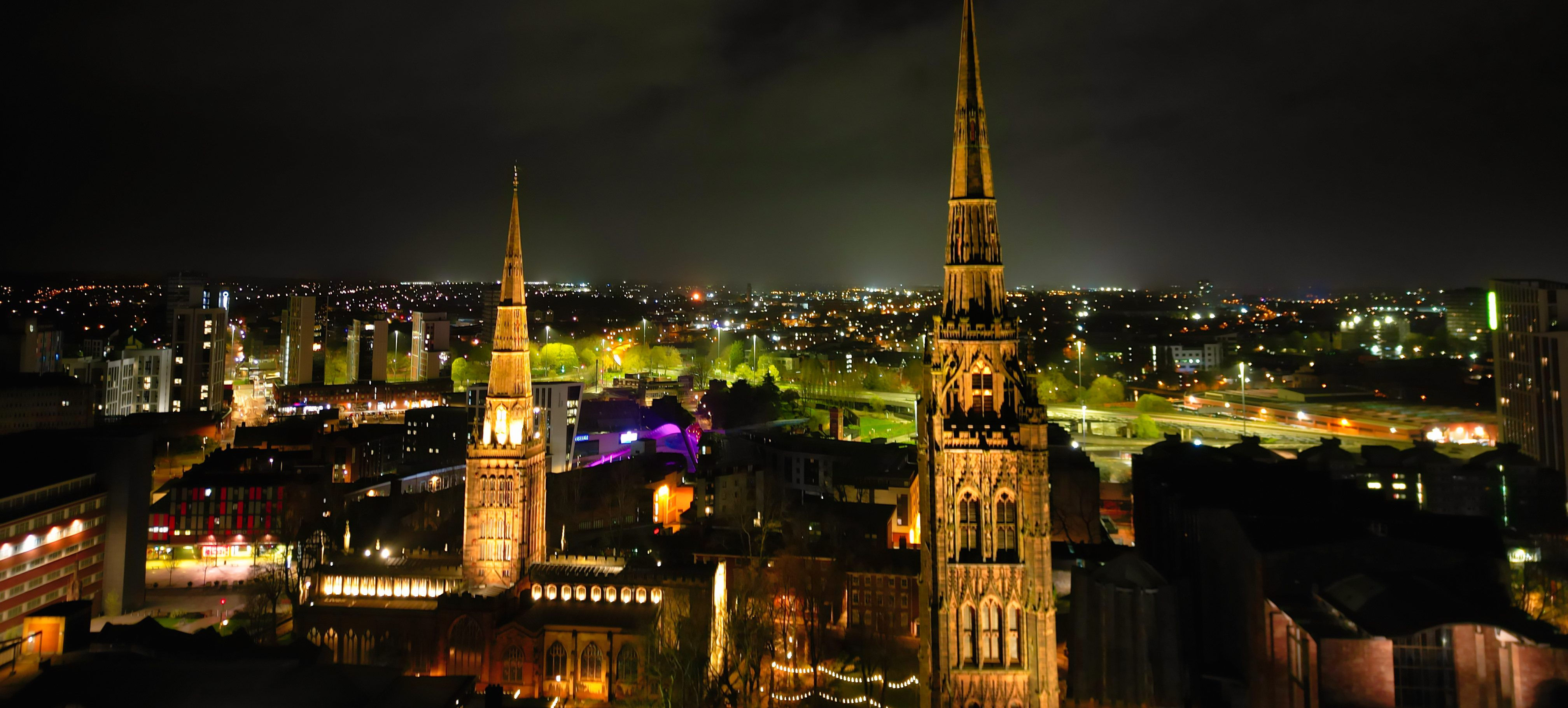 A night view of Coventry's cityscape reveals two illuminated church spires surrounded by urban buildings. The skyline, dotted with lights, creates a vibrant glow against the dark sky, unveiling hidden gems that students and locals alike cherish in their evening strolls.