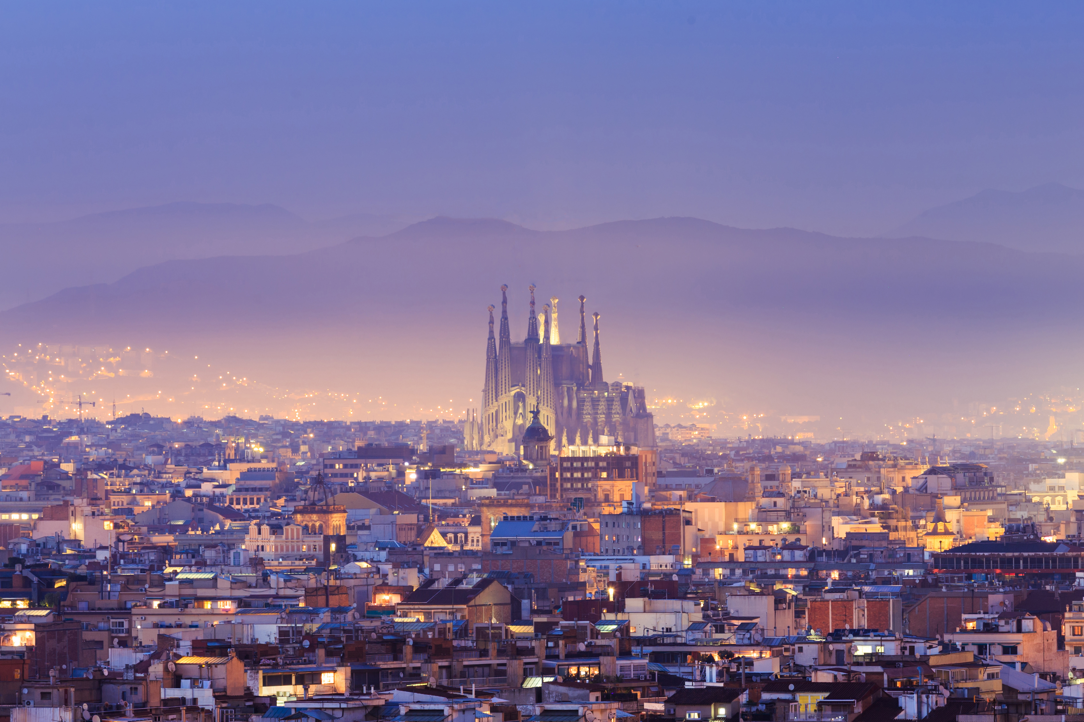 A panoramic view of Barcelona at dusk reveals the illuminated Sagrada Familia cathedral, sparkling in the center. The city lights twinkle under a hazy sky, with mountain outlines in the background—a perfect setting for those looking to study in Barcelona, home to some of the best universities.