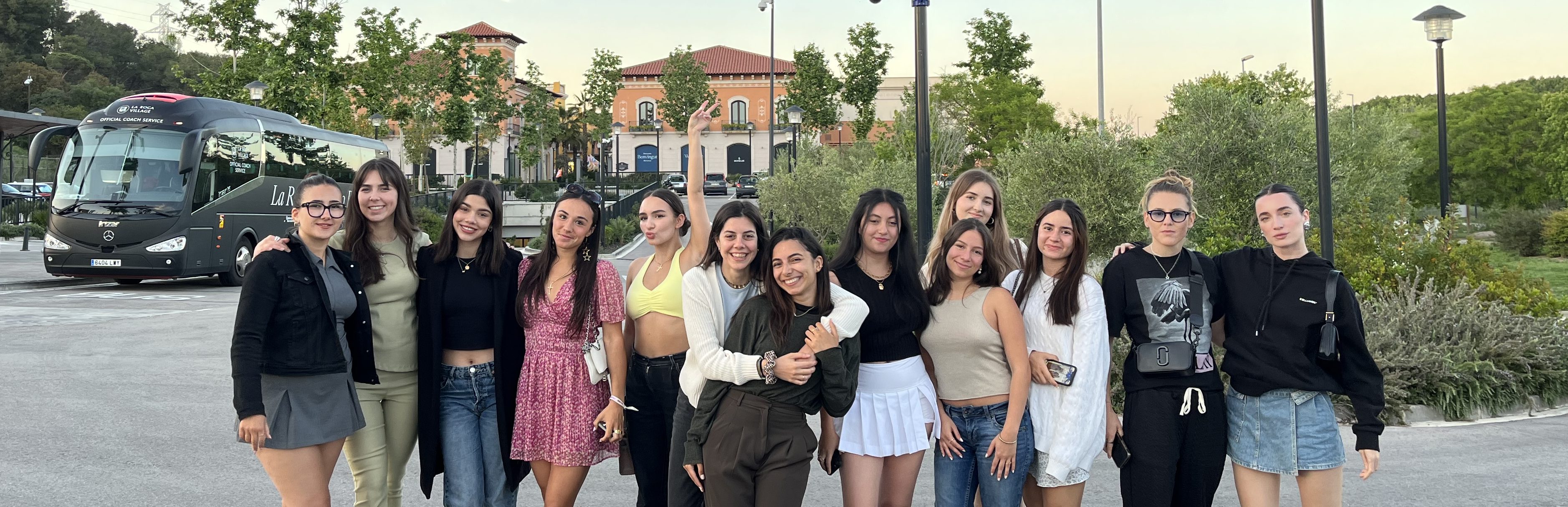 A group of fourteen international students stands together in an outdoor parking area, posing for a photo. They are dressed casually, with a mix of skirts, shorts, and dresses. A bus and trees are visible in the background.
