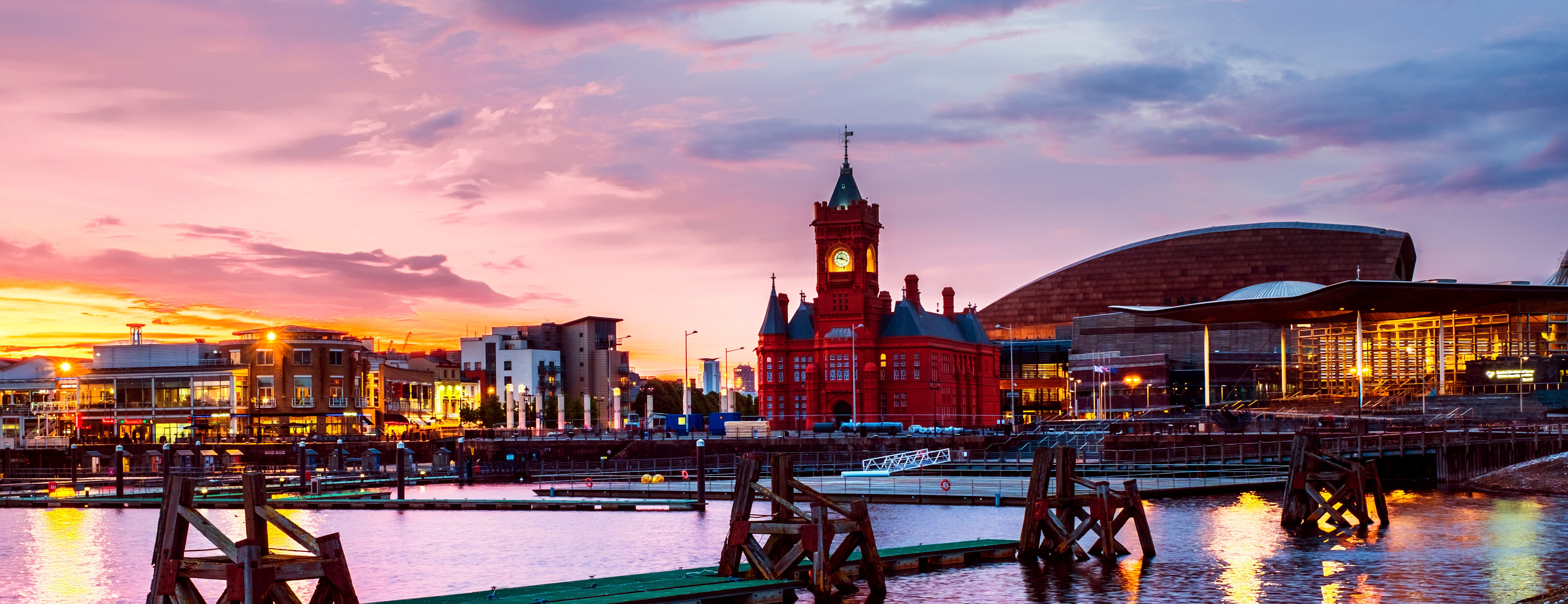 Panoramic view of Cardiff Bay at sunset, featuring the iconic red-brick Pierhead Building with a clock tower, the modern Wales Millennium Centre, and moored boats on the water reflecting vibrant hues. Nearby, lively bars invite students to unwind after a day of exploration.