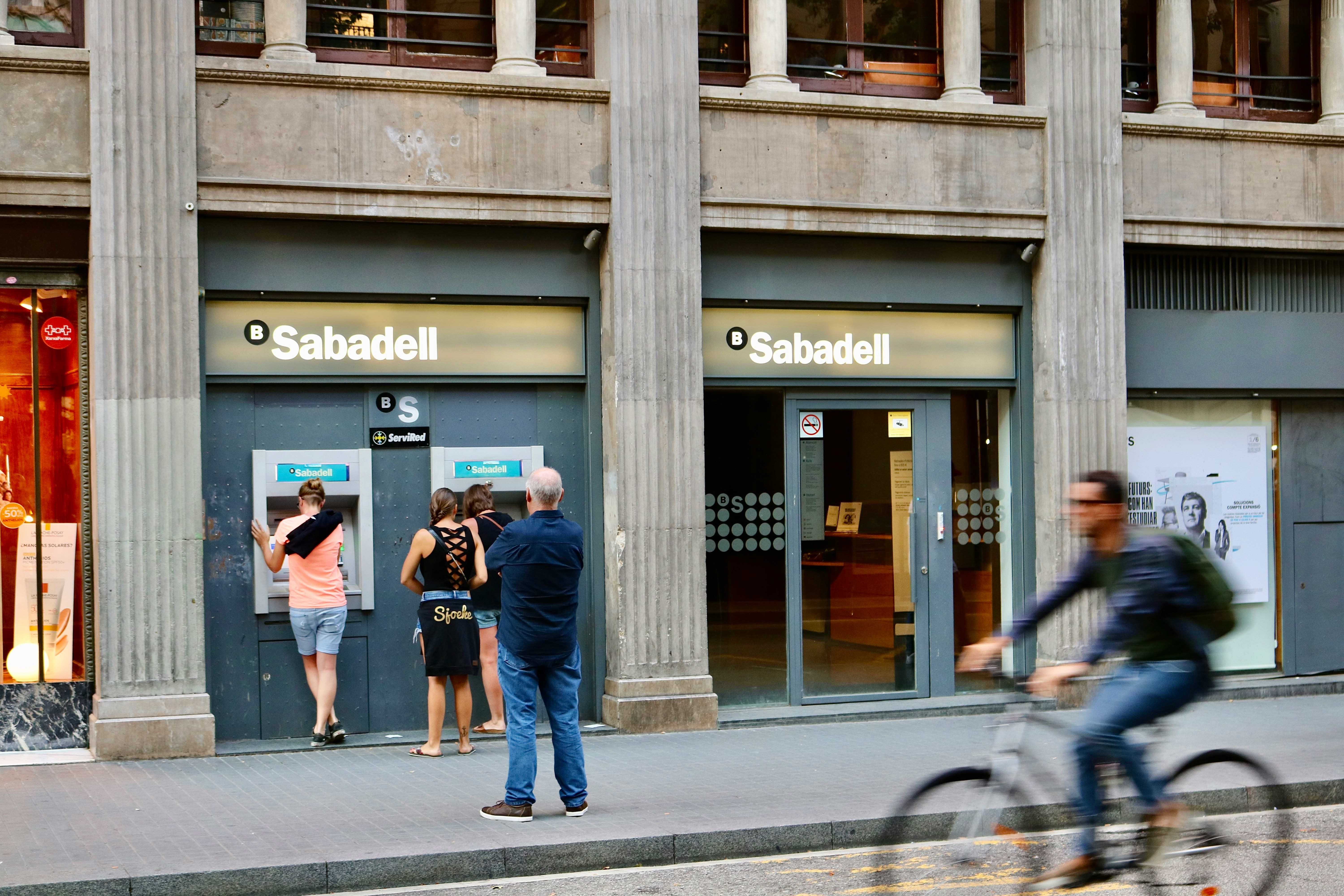 People standing at ATMs outside a Sabadell bank in Spain, with a cyclist passing by on the street. An international student waits patiently, perhaps ready to open a bank account. The building boasts tall columns and a modern facade, encapsulating the vibrant city life.