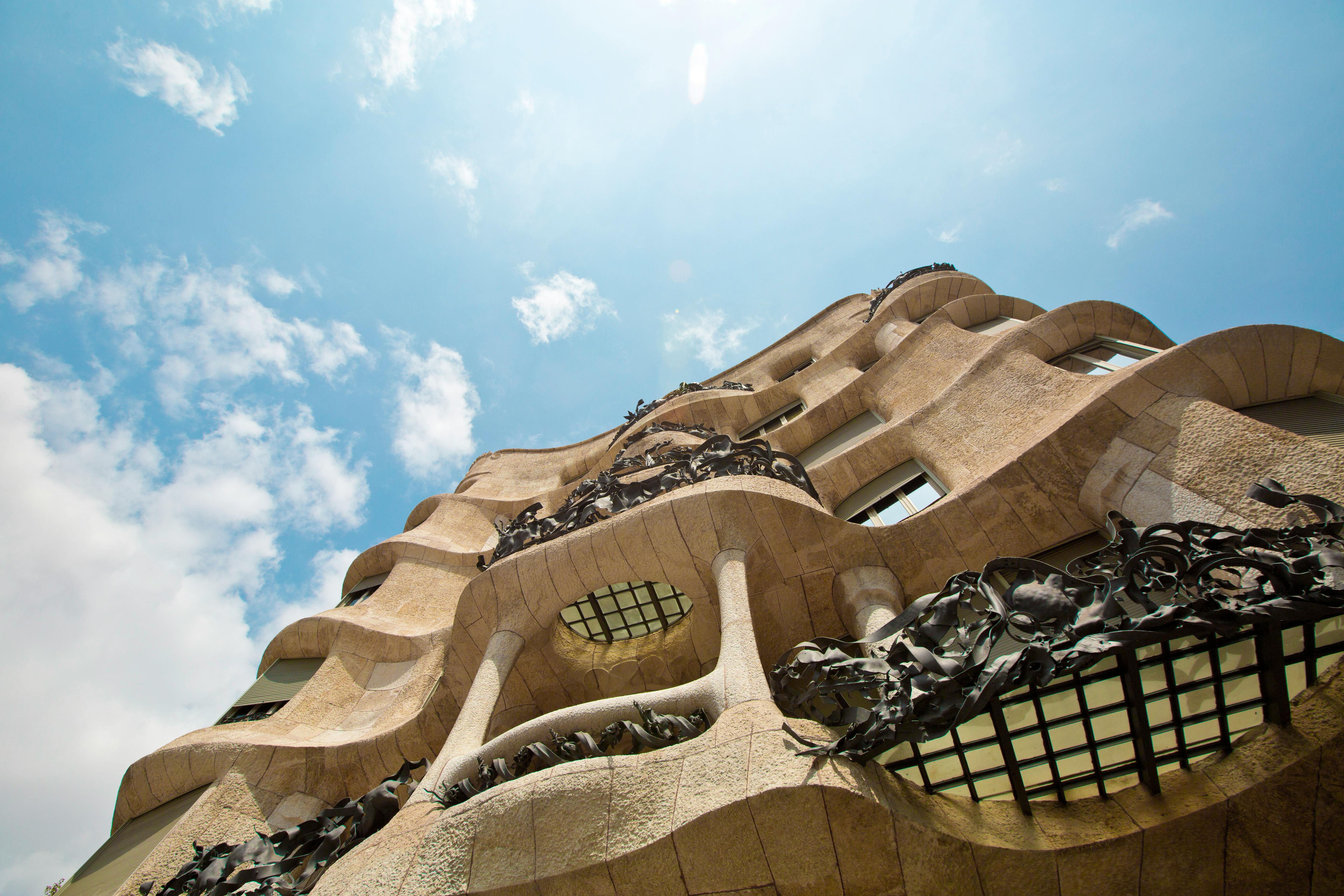 Upward view of a distinctive, wavy stone building with ornate ironwork balconies against a blue sky with scattered clouds. The architecture features curving lines and textured surfaces, reminiscent of the artistic flair found in Vita Student Barcelona designs.