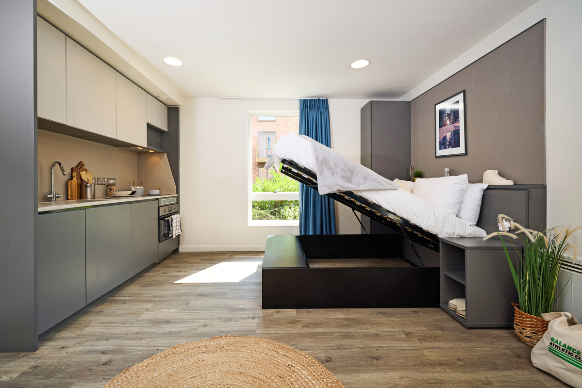 A modern studio apartment with a compact kitchen on the left, featuring gray cabinets and stainless steel appliances. A Murphy bed is partially lifted on the right, with storage underneath. The room has a wooden floor, a window, and a blue curtain.