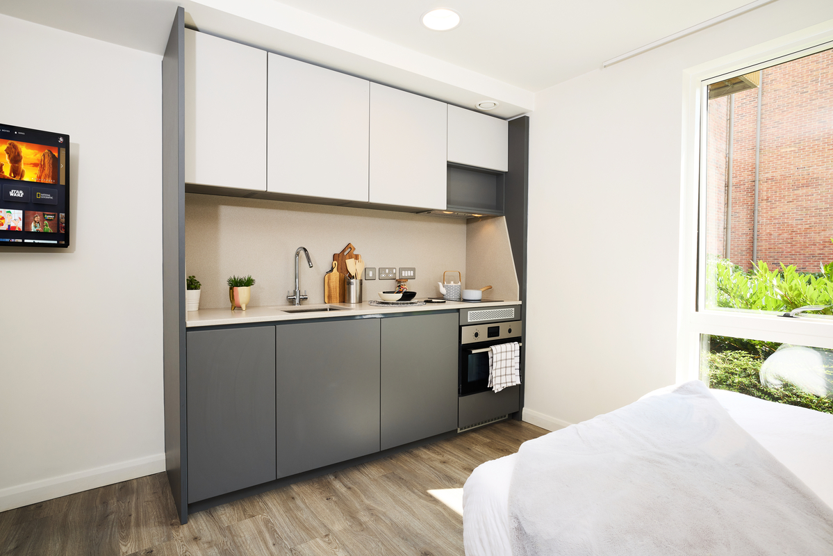 A modern, compact kitchen in a small apartment with sleek gray and white cabinets, a built-in oven, and a small sink. The room has a large window with a partial view of greenery, and a wall-mounted TV to the left. A bed is partially visible.