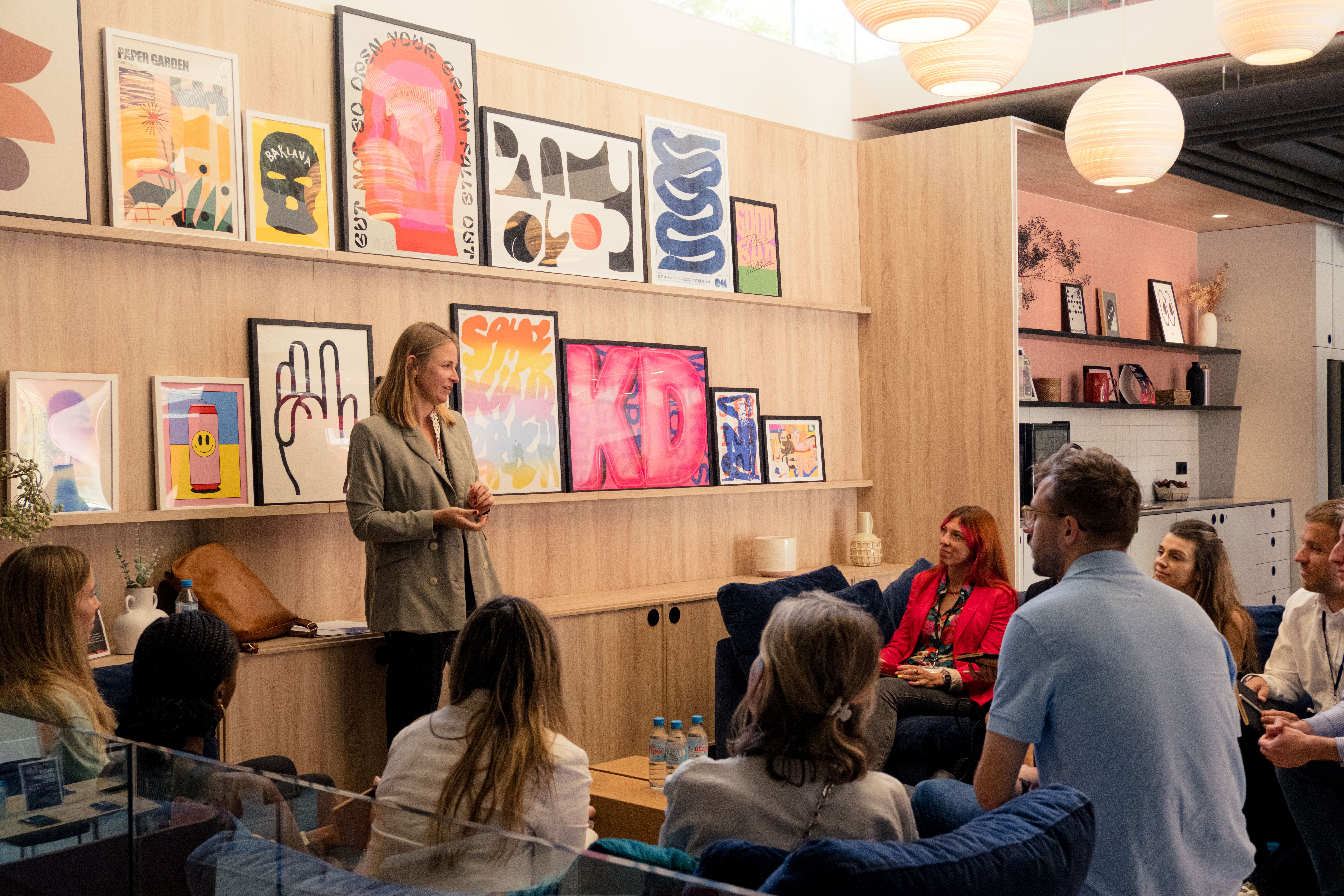 A woman stands and speaks to a seated group in a modern office space, giving them a head start on their careers. Artworks in various styles cover the wooden walls. The group listens attentively, with a bright and cozy atmosphere highlighted by hanging spherical lights.