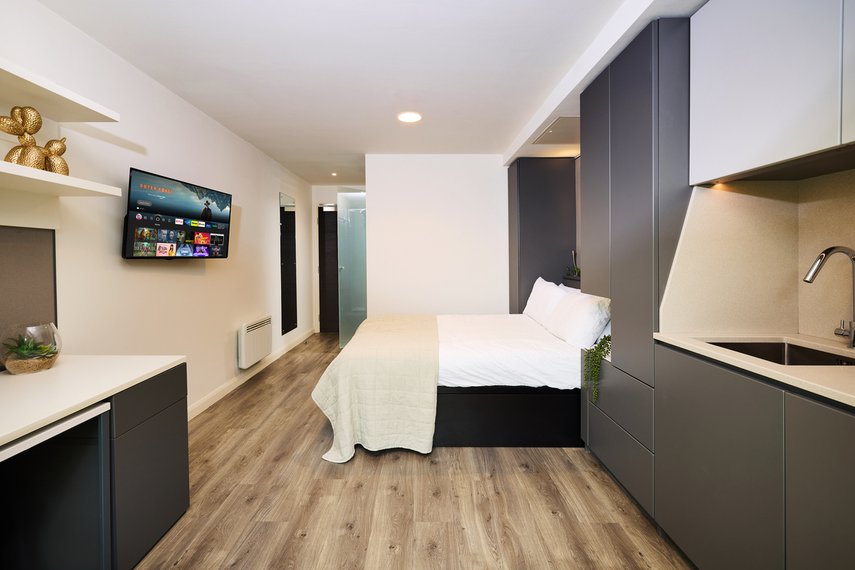 Modern studio apartment with a kitchenette, gray cabinets, and light wood flooring. A bed is in the center, facing a wall-mounted TV. A small workspace is beside the bed. The room is well-lit with a minimalist design.