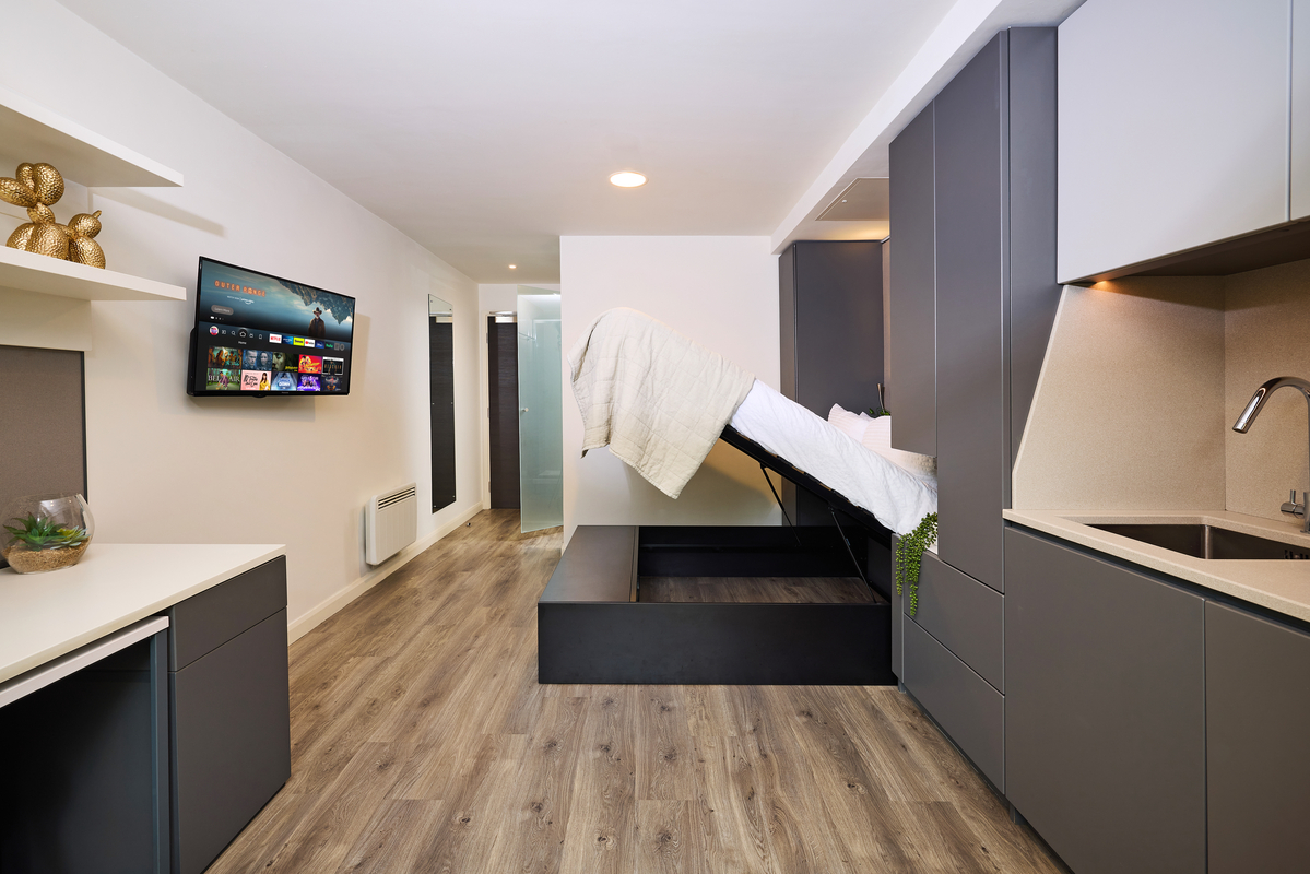 Modern studio apartment with a pull-down bed partially folded up against a gray wall. The room features wood flooring, a small kitchenette with a sink, a wall-mounted TV, and minimalistic decor, including shelves with decorative items.