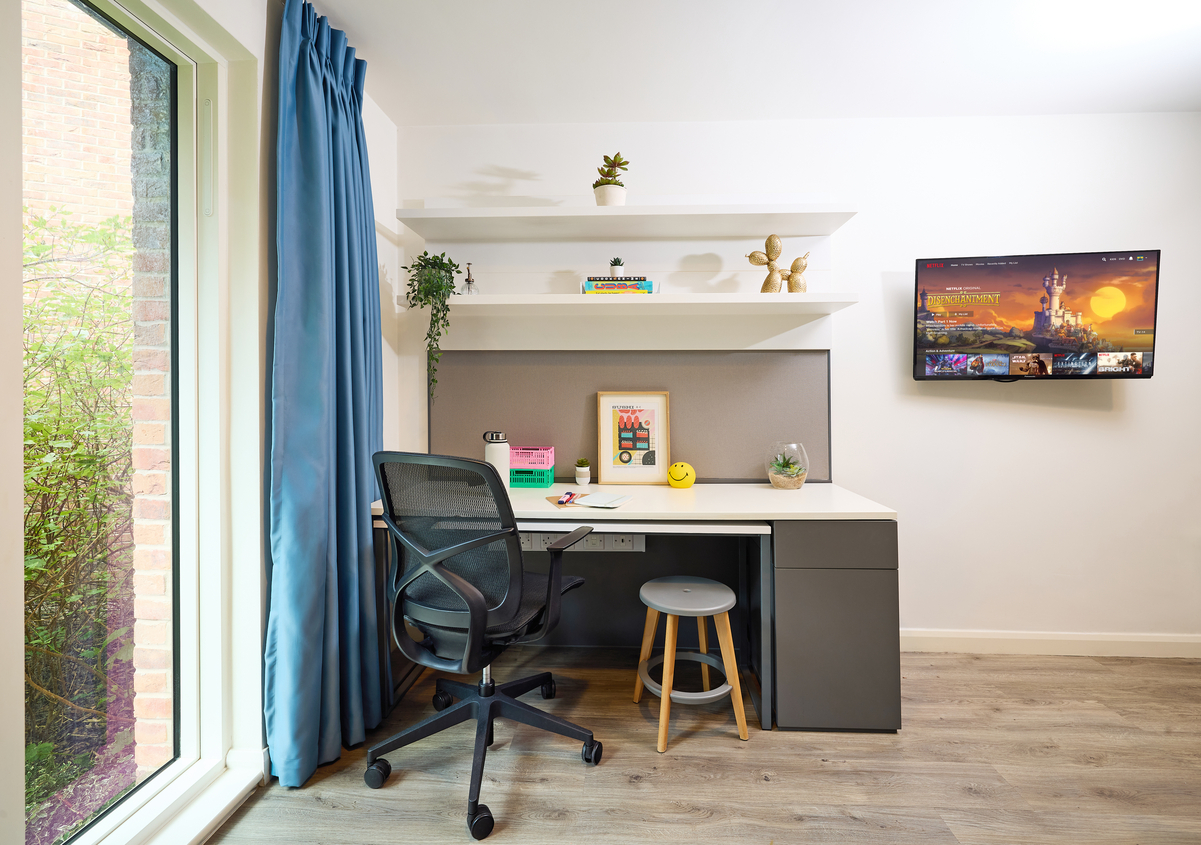A modern study area with a desk, chair, and small stool. Shelves hold plants and decor. A window with blue curtains is on the left, allowing natural light. A TV hangs on the opposite wall. Light wood flooring complements the bright, minimalist design.