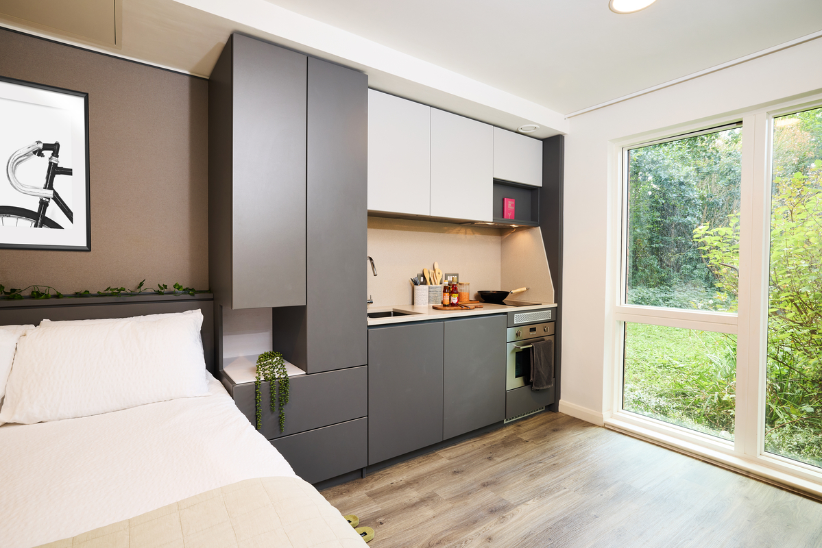 A compact, modern studio apartment featuring a small kitchen with dark cabinets, a sink, stove, and oven. A bed with white linens is adjacent, and a large window provides a view of green foliage outside. A framed bicycle print decorates the wall.