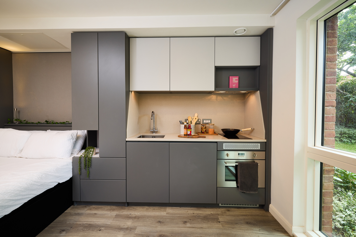Modern kitchenette with gray cabinets, a small sink, stove, and oven. Wooden utensils and decor are on the countertop. A bed is partially visible on the left, and a window on the right reveals a garden view. Light wood flooring completes the space.