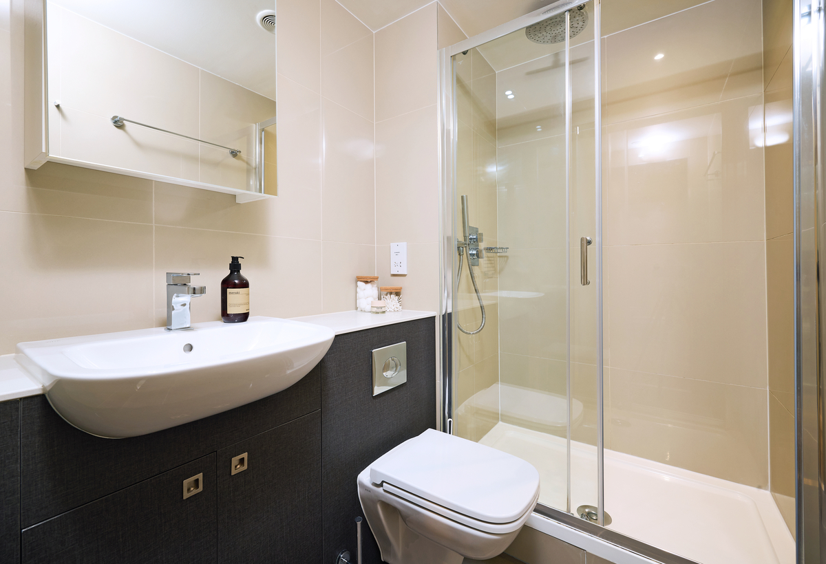 Modern bathroom with a white sink, soap dispenser, and a mirror cabinet above. A toilet is positioned beside a glass-enclosed shower. Beige tiled walls and a sleek design create a clean and minimalist aesthetic.