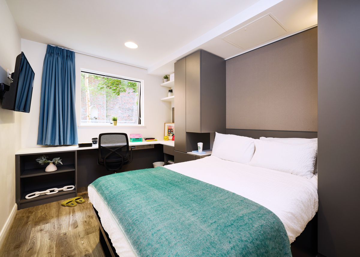 A modern, compact bedroom features a neatly made double bed with white bedding and a teal blanket. There's a desk with a chair, a TV mounted on the wall, shelves, and a window with blue curtains letting in natural light.