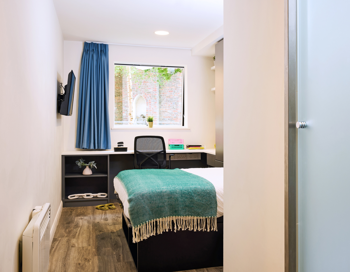 A minimalist bedroom with a single bed, blue blanket, and white pillow. There's a small desk with a black chair, a window with a view of a brick wall, and a mounted TV. The floor is wooden, and the room is brightly lit.