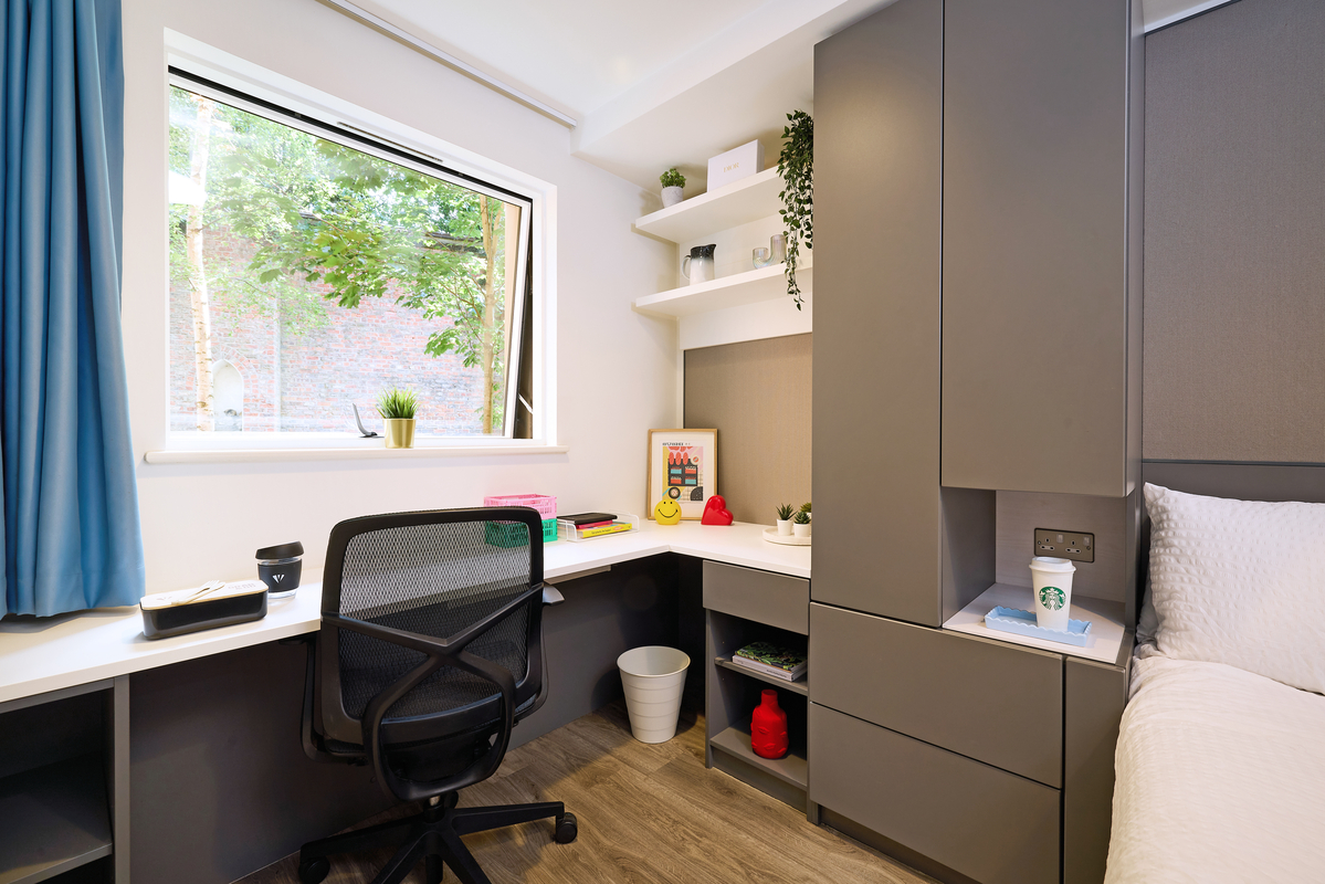 A cozy study room with a desk and chair by a large window with blue curtains. Shelves hold decorative items and a hanging plant. A bed with white bedding is next to a cupboard. Natural light brightens the room, and a small plant adds a touch of greenery.