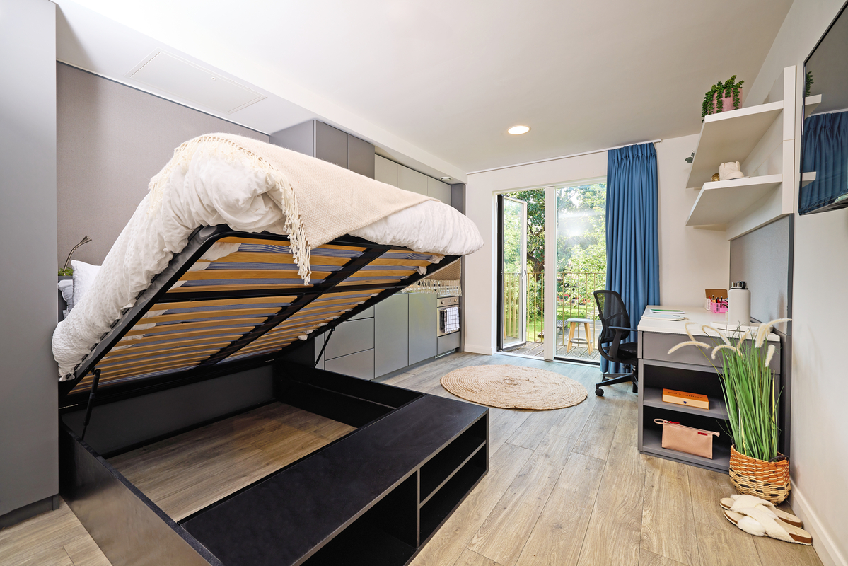 Modern studio apartment with a lifted bed revealing storage space beneath. There's a small kitchen, a round rug, a desk with a black and white chair, and a wall-mounted TV. Glass doors lead to a balcony with a view of trees.