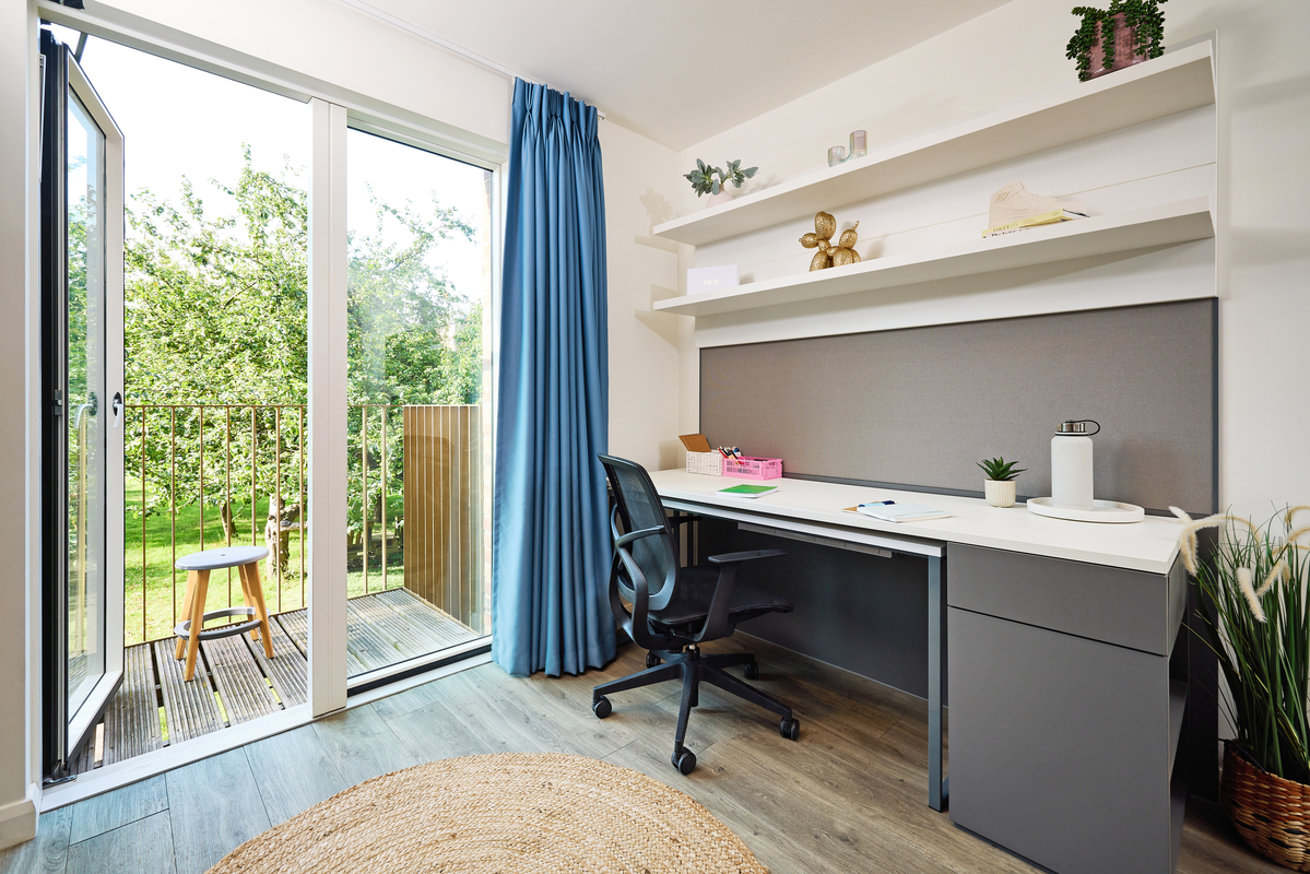 A modern home office with a desk and swivel chair facing a large window. The room has blue curtains, a wooden floor, and a round rug. Shelves above the desk hold decor items. A balcony with a view of greenery is visible outside.