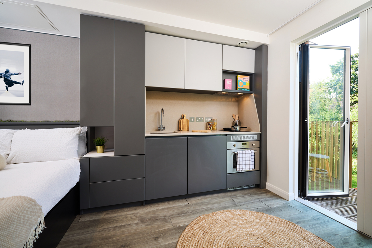 A modern studio apartment features a compact kitchen with gray cabinets, an oven, and a kettle. A bed is adjacent, and a round jute rug lies on wooden flooring. A door opens to a balcony with greenery outside. A framed picture decorates the wall.