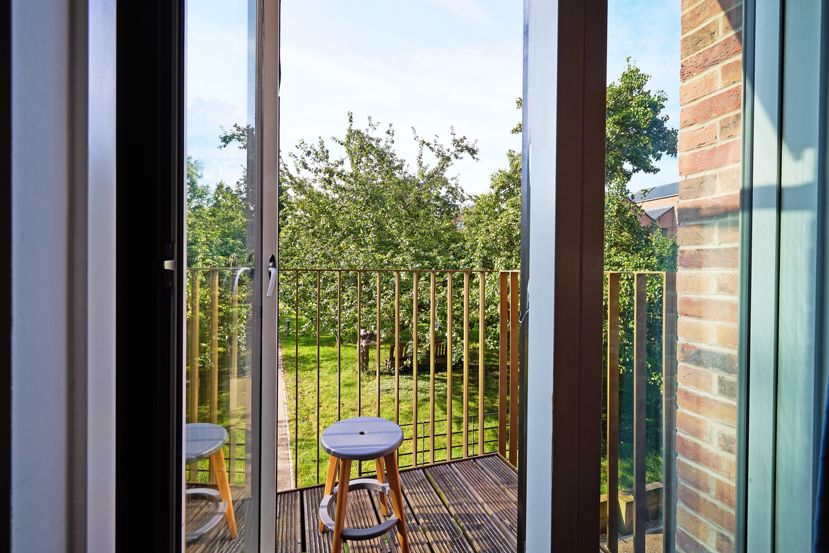 Open glass door leading to a small balcony with a wooden stool. The balcony overlooks a lush, green landscape with trees under a clear sky. A brick wall is partially visible on the right side.