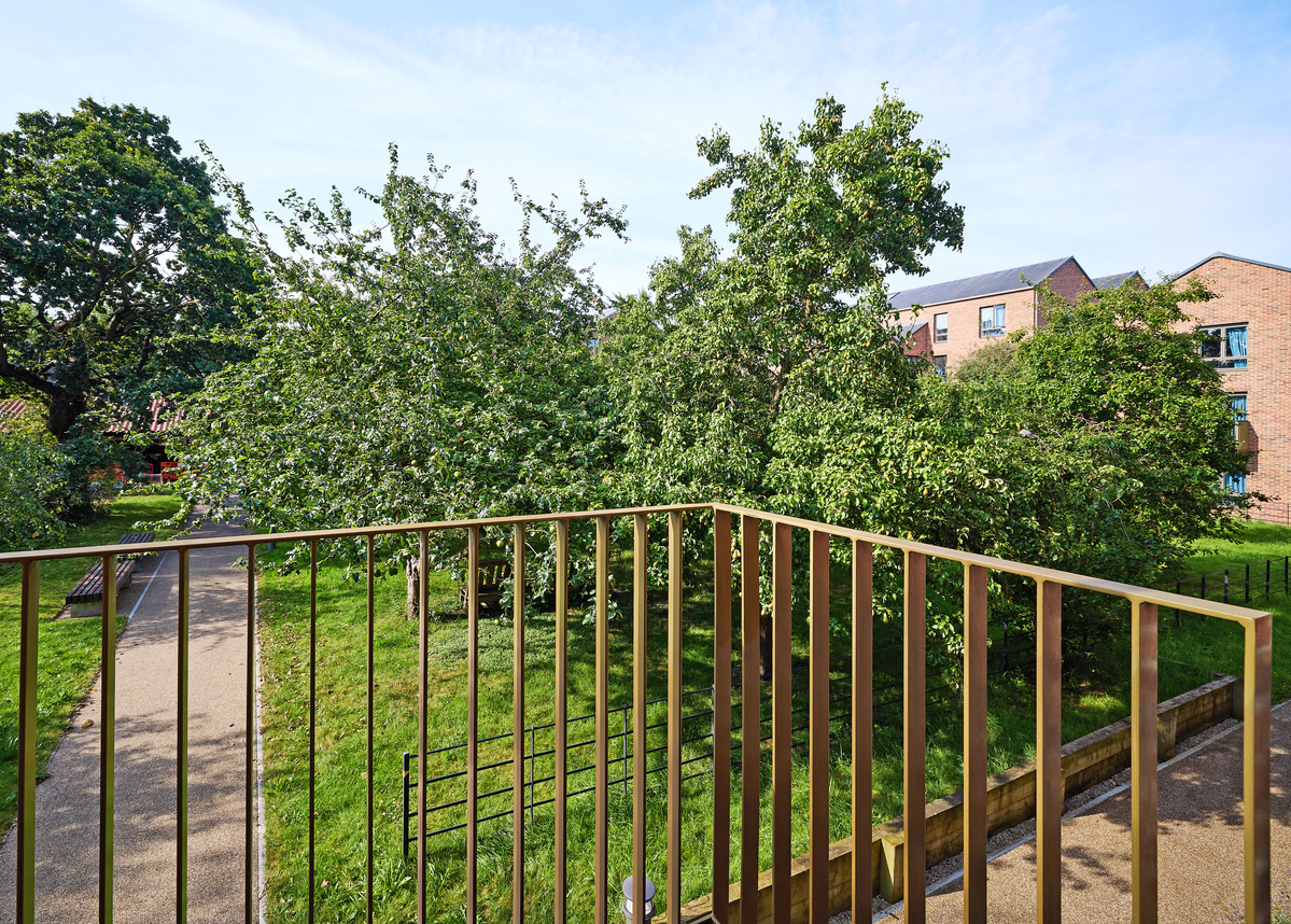 View from a balcony overlooking a park with lush green trees and a pathway. A modern brick building is visible in the background against a clear blue sky.