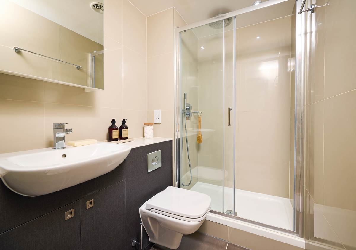 Modern bathroom featuring a wall-mounted sink, toilet, and a glass-enclosed shower with chrome fixtures. Cream and gray tiles cover the walls and floor. A mirror and toiletries are on the sink counter, with a towel hanging inside the shower.