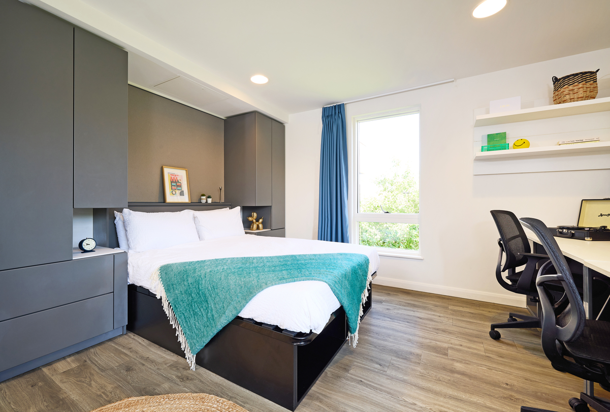 A modern bedroom with a neatly made bed, teal blanket, and gray cabinetry. A window with blue curtains brightens the room. There's a desk with a chair and shelves holding decor items. The hardwood floor adds warmth to the space.