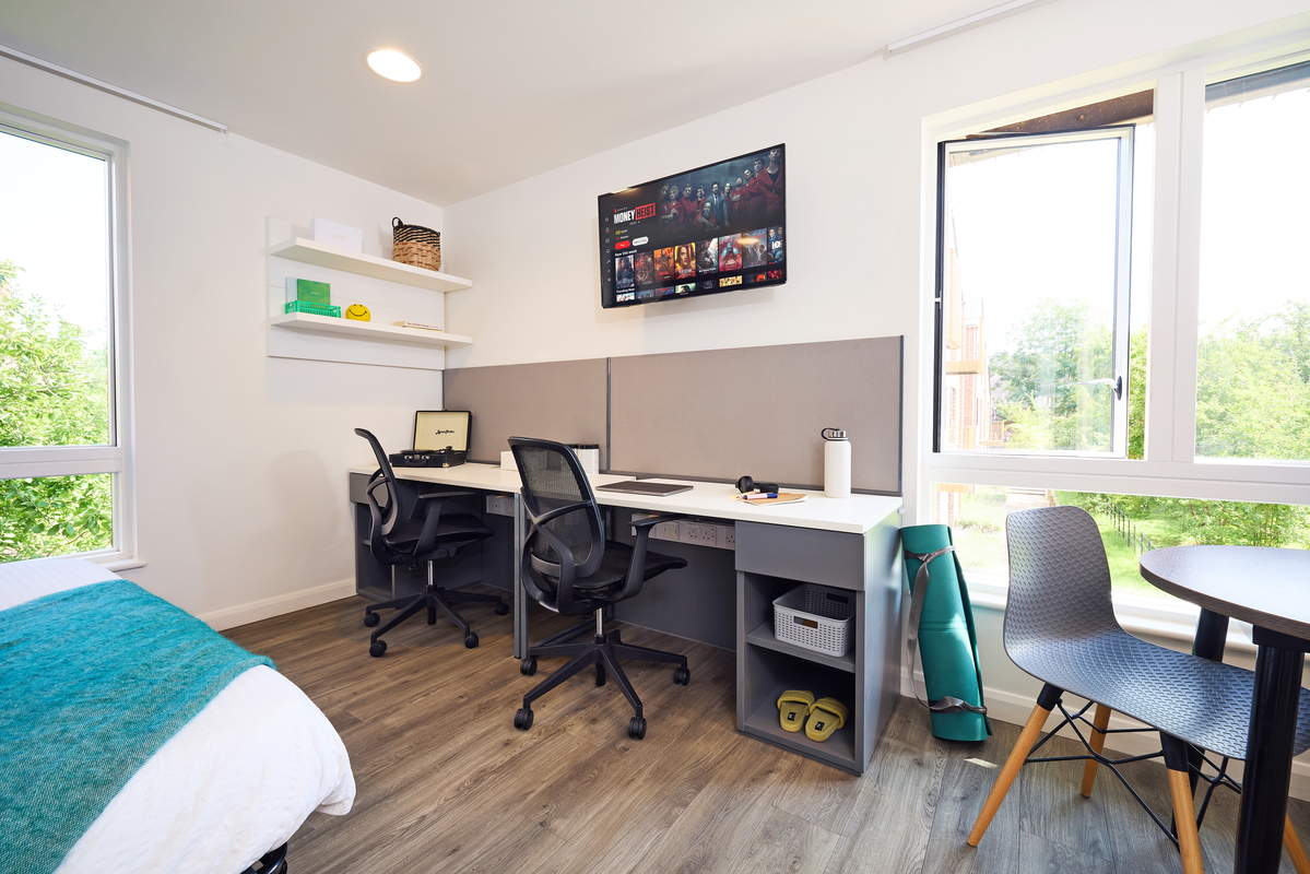 Modern bedroom with two desks, office chairs, and a wall-mounted TV displaying Netflix. Open laptop on one desk. Shelving with decor items above. Nearby bed with a teal blanket. Large windows with outdoor view and a small table with chairs complete the room.