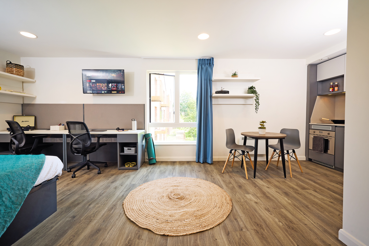 Modern studio apartment interior with a wooden floor. It features a bed, two desks with chairs, wall-mounted TV, and shelves. A round dining table with chairs is near a window with a blue curtain. Kitchenette with appliances is on the right side.