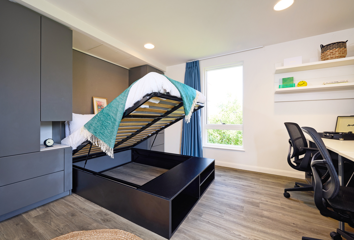 A modern room with a raised bed revealing storage underneath. The space features a desk with chairs, shelves with books and decor, and a large window with blue curtains. The floor is wooden, and the walls are white and gray.