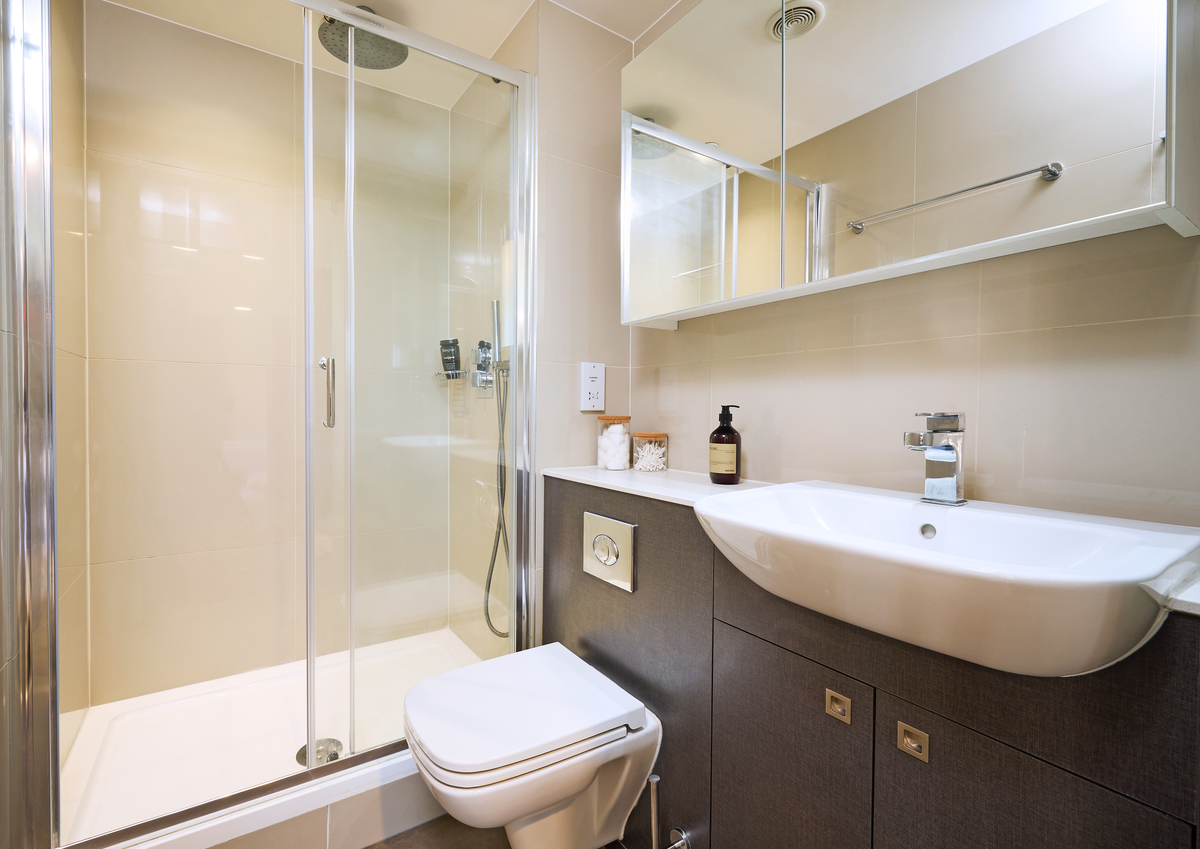 Modern bathroom with a glass-enclosed shower, a wall-mounted sink, and a toilet. The vanity has a dark wood finish with storage space, and a mirror with a shelf above. The walls are light beige with recessed lighting. Soap dispenser on the counter.