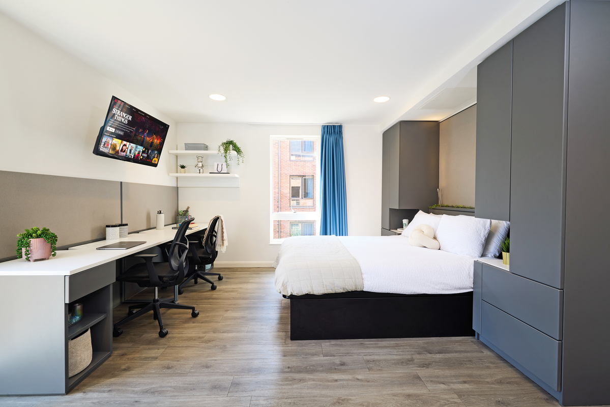 Modern bedroom with a large bed, blue curtains, and wood flooring. A wall-mounted TV is above a long desk with two chairs and computer monitors. Shelves with plants and decorations are above the desk. Built-in wardrobes are on the right.